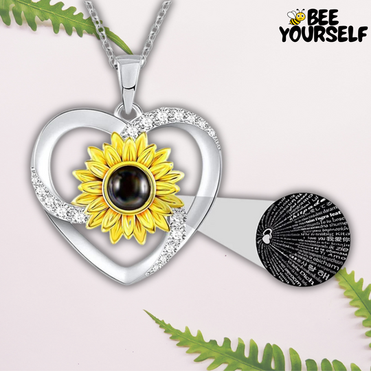 Collier pendentif en forme de cœur avec un tournesol central, entouré de cristaux scintillants. Au centre du tournesol, une pierre noire avec l'inscription 'Je t'aime' en 100 langues différentes, sur fond argenté. Le collier est de la marque Bee Yourself, posé sur un fond clair avec des feuilles de fougère décoratives.